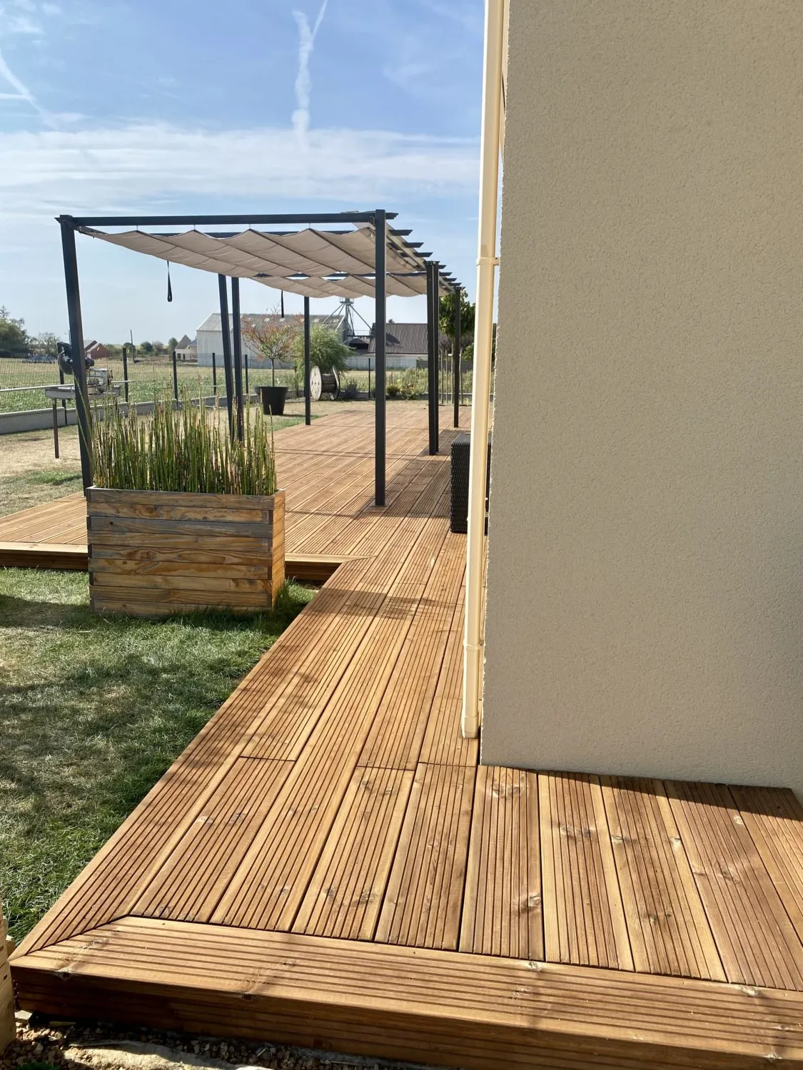 Terrasse en lames de bois clair posées en longueur, coin salon avec table basse en bois, vue sur un jardin et un ciel nuageux