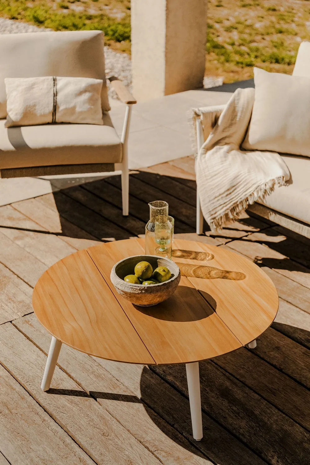 Table basse en bois brut posée sur une terrasse en pierre face à la mer, avec deux tasses en grès et un lin froissé